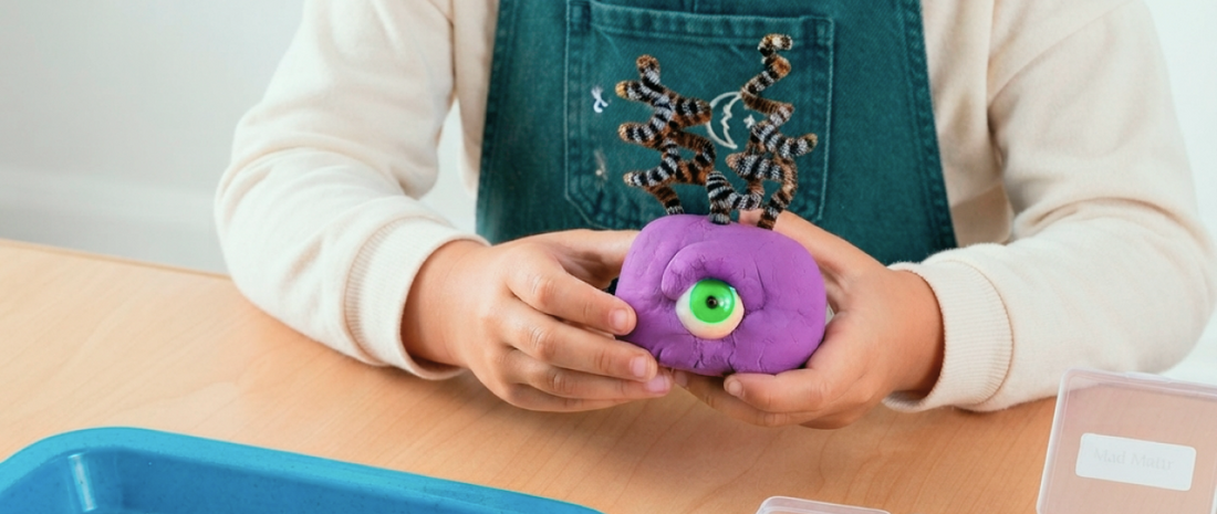  Child's hands molding a purple Mad Mattr monster with googly eyes and pipe-cleaner arms — mess-free Halloween craft for kids