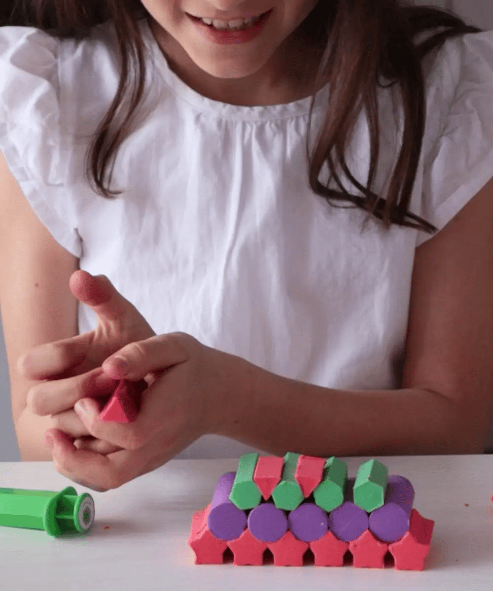 A child building a colorful tower with MadMattr sensory dough. The image shows the dough's ability to hold firm shapes like stars, cylinders, and triangles in vibrant shades of pink, purple, and green.